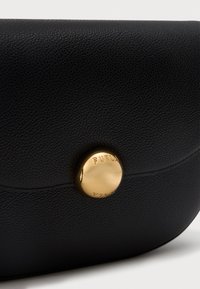 Close-up laukku mustasta teksturoidusta nahasta, jossa on pyöreä kiiltävä kultainen napin kiinnitys, jossa on kaiverrus "FURLA".