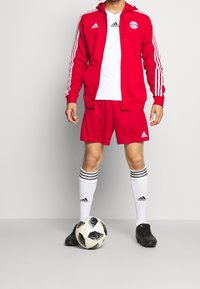 Survêtements sportifs Adidas rouge avec des rayures blanches, T-shirt blanc et shorts assortis, associés à des crampons noirs et des chaussettes blanches montantes.