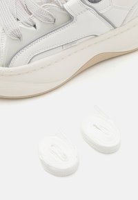 Baskets blanches avec une tige en cuir texturé et en mesh, présentant des accents gris. Deux rouleaux de lacets blancs positionnés sous la chaussure.
