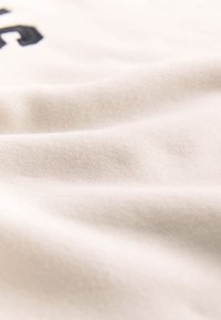 Tissu doux de couleur crème avec des plis subtils et une vue partielle d’un texte imprimé en noir dans le coin supérieur gauche.