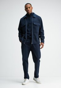 Marineblauwe zip-up jas met twee borstzakken, gecombineerd met een gestructureerde trui en bijpassende broek, aangevuld met witte sneakers.