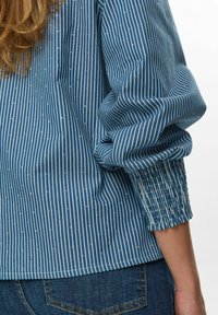 Zils svītrots tops ar elastīgiem piedurknēm, piedāvājot plānas baltas vertikālās līnijas un mazus metāla akcentus. Audums izskatās viegls un tekstūrēts.