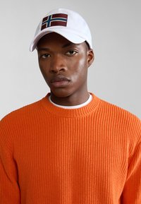 Cappello bianco con design di una bandiera maroon e blu, abbinato a un maglione arancione a coste texturizzato. Sfondo minimalista.