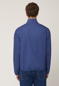 Marinblå ofodrad jacka med ståkrage, långa ärmar, elastisk midja och slät tygtextur, bärs över denimjeans.