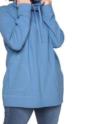 Personne portant un sweat à capuche bleu texturé avec des poches avant, tirant sur les cordons de la capuche, associée à un jean bleu sur un fond blanc.
