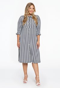 Schwarz-weiße Knopf-Up-Kleid mit gemusterten Dreiecken, Dreiviertelärmeln, gerafften Manschetten und einem leicht ausgestellten Saum. Akzentuiert mit runden Knöpfen.
