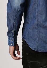 Blå denimskjorta med nedknäppt krage, långa ärmar och tre vita knappar vid manschetten. Visar en avslappnad passform och texturerad tyg.