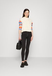 Suéter creme de malha trançada com mangas listradas multicoloridas, jeans skinny pretos, mocassins pretos robustos e uma bolsa preta.