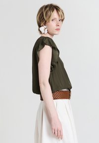 Top corto a maniche corte color verde oliva, con texture e bordi arricciati, abbinato a una cintura intrecciata marrone, indossato con una gonna bianca a pieghe.