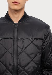 Fekete steppelt bomberdzseki bordázott gallérral, középső cipzárral és vízszintes díszcsíkokkal a mellkason. Puha, sima textúra.