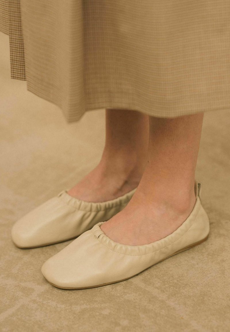 Smėlio spalvos odiniai balerinai su apvaliu priekiniu kraštu, elastingu apvadu ir lygia tekstūra, ilsintysis ant šviesios spalvos paviršiaus.