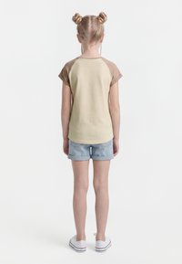 Barn med blondt hår i to boller, iført beige og brun t-skjorte med korte ermer, lyse denimshorts og hvite joggesko, står med ryggen til.