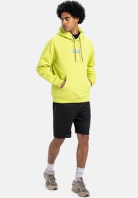 Bright lime green hoodie with a kangaroo pocket and drawstring, paired with black shorts. Features graphic embroidery on the chest.