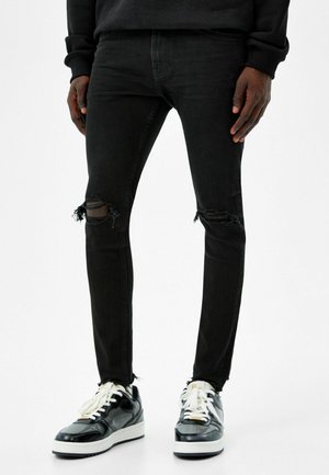 Personne portant un jean skinny noir déchiré et des baskets noires et blanches, debout devant un fond uni.
