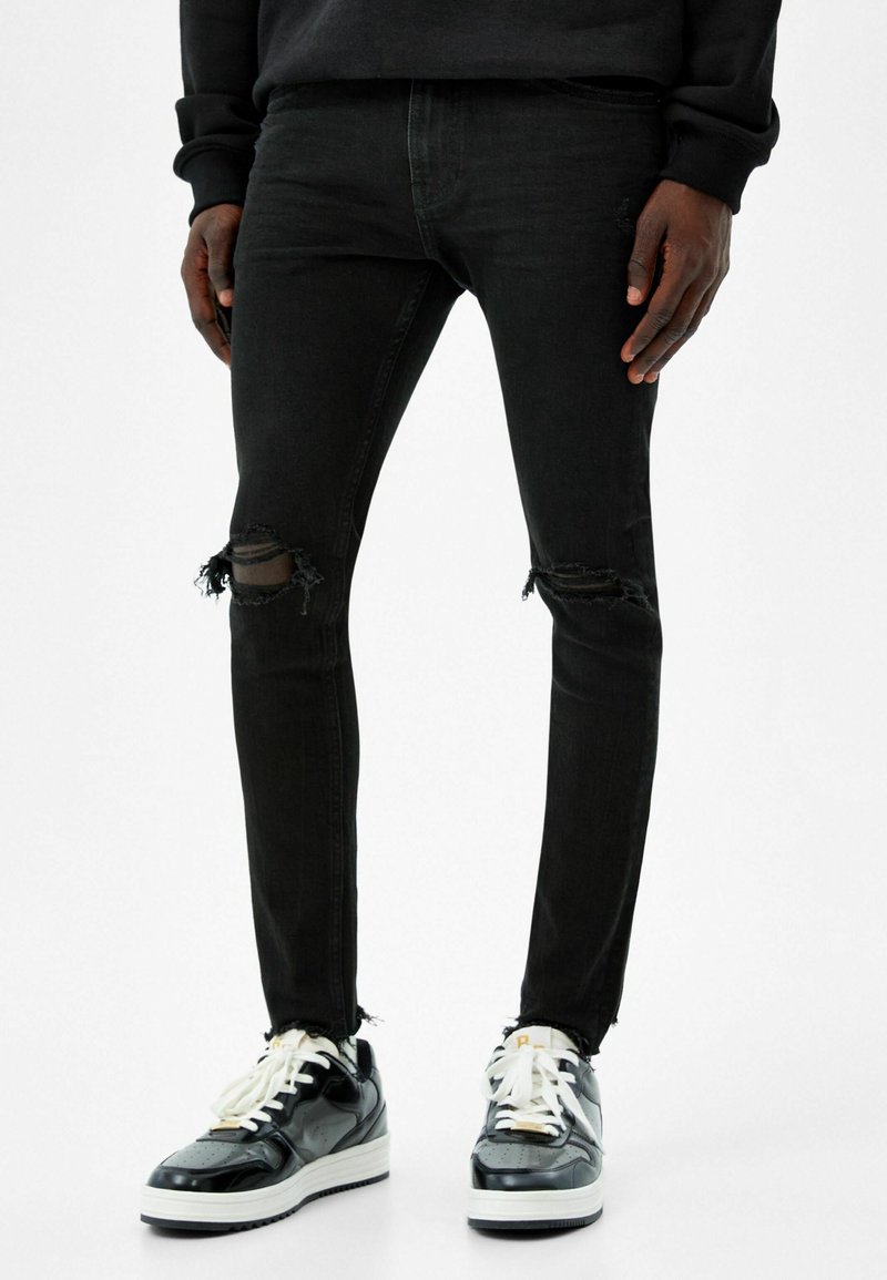 Personne portant un jean skinny noir déchiré et des baskets noires et blanches, debout devant un fond uni.
