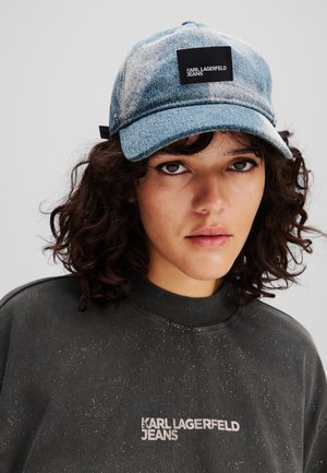 Gorra de béisbol de denim con un patrón azul y blanco, que presenta un parche de logo negro. Acompañada de un sudadera oscura con una textura brillante.