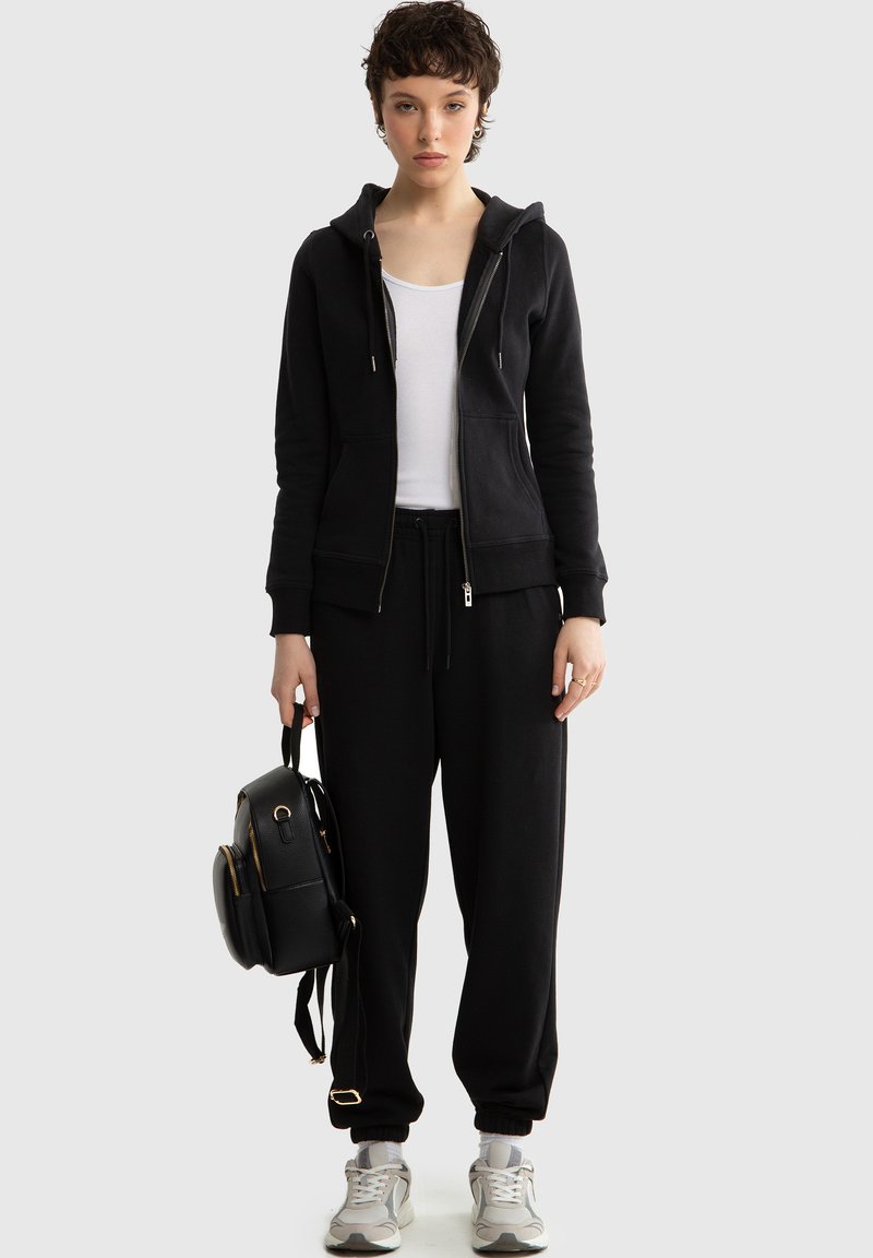 Vrouw met kort haar, gekleed in een zwarte hoodie met rits, zwarte joggingbroek, wit hemdje en grijze sneakers, houdt een kleine zwarte rugzak naast zich vast.