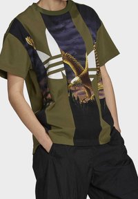 Olivgrön och svart kortärmad t-shirt med en stor vit Adidas-logotyp och en framträdande örn-grafik på framsidan.