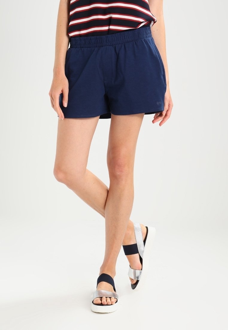 Osoba oblečená v námořnických šortkách na elastický pas, pruhovaném topu bez rukávů a stříbrno-černých sandálech na pásku, stojící s překříženýma nohama.