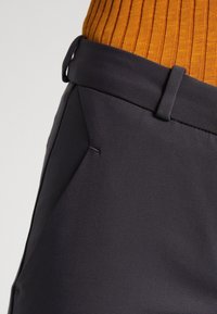 Pantaloni neri su misura realizzati in tessuto liscio, con fronte piatto, una tasca singola e passanti per la cintura. Maglia arancione a coste, parzialmente visibile.