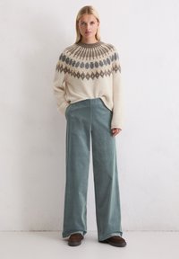 Sweter z dzianiny w kolorze kremowym z wzorzystym karczkiem w odcieniach szaro-brązowych, połączony z szerokimi spodniami typu corduroy w kolorze zielonym oraz brązowymi butami wsuwanymi.