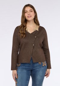 Brun texturerad cardigan med V-ringning och guldfärgade knappar, kombinerad med en beige skjorta med knappar under, bärs tillsammans med blå broderade jeans.