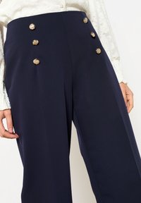 Pantaloni navy a vita alta con una texture liscia, caratterizzati da quattro bottoni decorativi color oro sul davanti per un tocco in più.