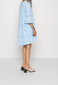 Blå bomullsklänning med hålmönster, lager på lager volanger och trekvartslånga klockärmar, kombinerad med öppna tofflor med tjock klack.