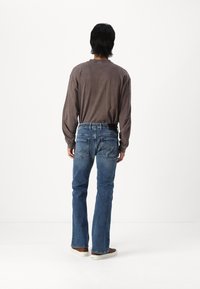 Camisa marrón de manga larga, vaqueros azules con un acabado desvaído y zapatillas marrones. Los vaqueros presentan una costura distintiva en los bolsillos traseros.