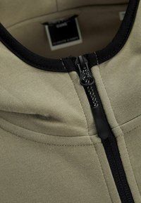 Haut de survêtement zippé vert olive avec une fermeture éclair et des finitions noires. Tissu lisse avec des détails de couture plate et un design minimaliste.