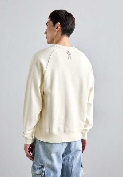 Man facing away wears cream sweatshirt with small astronaut graphic on upper back and light blue cargo jeans.