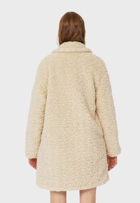 Frau mit langen braunen Haaren, die einen knie-langen, cremefarbenen, strukturierten Teddy-Mantel trägt, von hinten vor einem schlichten Hintergrund gezeigt.