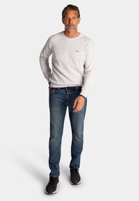 Middeljarige man met kort haar in een lichtgrijze trui, blauwe spijkerbroek, zwarte riem en zwarte sneakers, staand tegen een witte achtergrond.