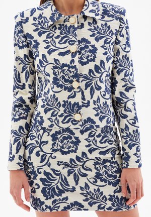 Weiß strukturiertes Jacken- und Rockset mit blauem Blumenmuster und weiß mit Gold besetzten Knöpfen, getragen von einer stehenden Person.