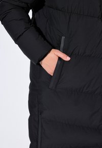 Černá péřová bunda s detailním pohledem na ruku v boční kapse, s prošívanou texturou a elegantními detaily.