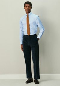 Svetlomodrá kockovaná košeľa, hnedá vzorovaná kravata, tmavomodré nohavice a hnedé kožené topánky, stojaci pred neutrálnym pozadím.