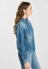 Woman in blue denim shirt and jeans standing in profile against a plain light background.