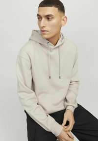 Light beige hoodie with drawstrings and front pocket. Smooth texture, relaxed fit, long sleeves, and ribbed cuffs, worn over black pants.