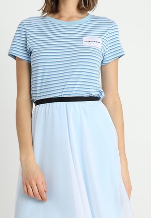 Nainen, jolla on sinivalkoraidallinen Calvin Klein Jeans -t-paita, joka on tungettu vaaleansiniseen hameeseen, jossa on musta vyö.