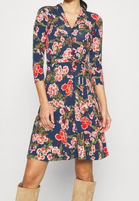 Kvinna i en marinblå wrapklänning med röd och vit blommig mönster, trekvartsärmar, en knytmidja och beige knähöga stövlar i mocka.