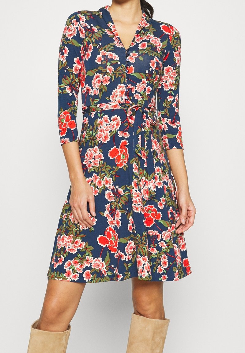 Kvinna i en marinblå wrapklänning med röd och vit blommig mönster, trekvartsärmar, en knytmidja och beige knähöga stövlar i mocka.