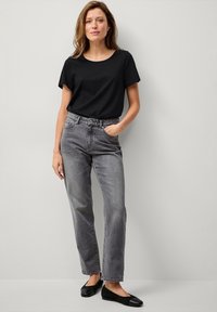 Schwarzes T-Shirt mit Rundhalsausschnitt kombiniert mit hochgeschnittenen grauen Jeans. Die Schuhe sind schwarze Ballerinas. Glatte Textur und lockere Passform mit Seitentaschen.
