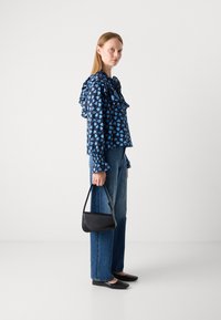 Blusa blu con motivo a cuori e colletto arricciato, abbinata a jeans azzurri e scarpe basse nere. Il modello tiene una piccola borsa nera.