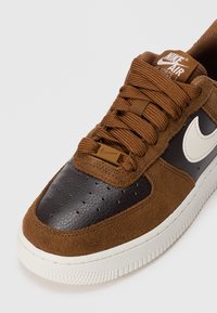 Smeđi suđeni i kožni tenisice s bijelim Nike Swoosh-om, teksturiranim prednjim dijelom i smešnim vezicama na bijeloj gumi.