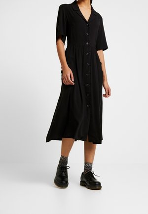 Vestido midi negro con botones en el frente, mangas cortas y un cuello. Tela suave con cintura definida y falda fluida. Combinado con zapatos negros y calcetines estampados.