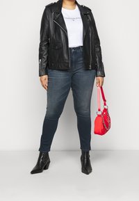 Chaqueta de moto de cuero negra, camiseta gráfica blanca, jeans skinny de lavado oscuro, botas de tacón negras, sosteniendo un pequeño bolso rojo con detalle de cadena.
