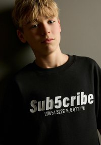 Sort sweatshirt med fed hvid tekst "Sub5cribe LDN 51.5229° N, 0.0777° W." Crew neck design med en afslappet pasform.