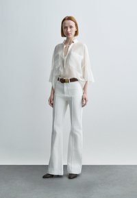 Mujer de pie sobre un suelo gris que lleva una blusa blanca transparente, pantalones acampanados blancos de cintura alta, cinturón marrón y zapatos oscuros puntiagudos, con fondo neutro.