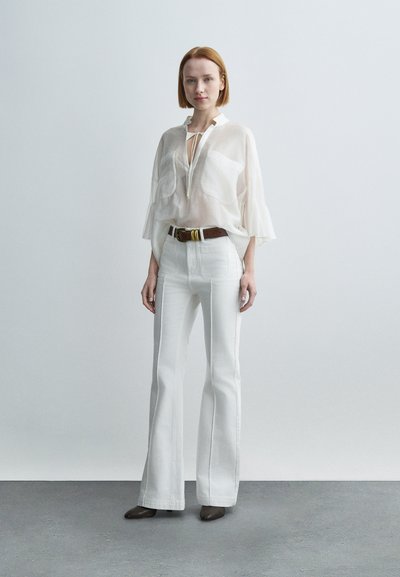 Γυναίκα στέκεται σε γκρι πάτωμα φορώντας διαφανές λευκό μπλουζάκι, λευκό παντελόνι με ψηλή μέση και καμπάνα, καφέ ζώνη και σκούρα μυτερά παπούτσια, ουδέτερο φόντο.