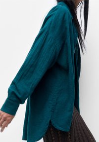 Chemise boutonnée bleu sarcelle en tissu texturé, avec manches longues, fentes latérales et coupe décontractée, associée à une jupe foncée à motifs.